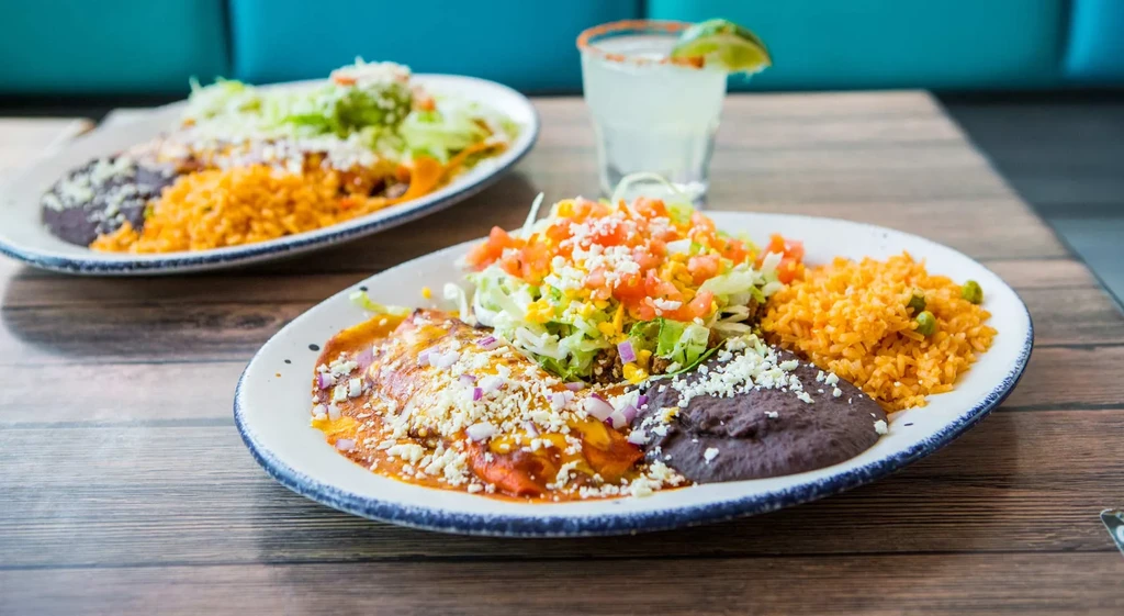Two plates of Mexican enchiladas, beans, rice, and salad on a table.