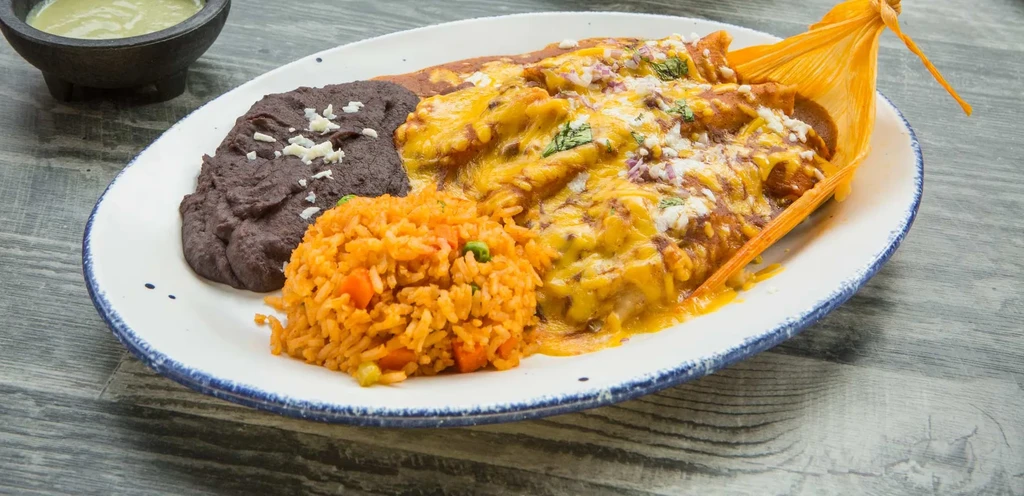 Plate of cheesy Mexican food with beans, rice, and a corn husk.