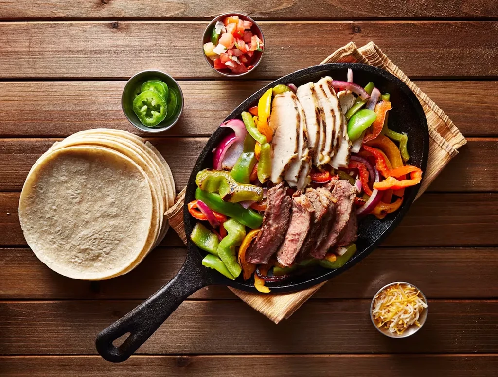 Chicken and steak fajitas in a skillet with tortillas and toppings.