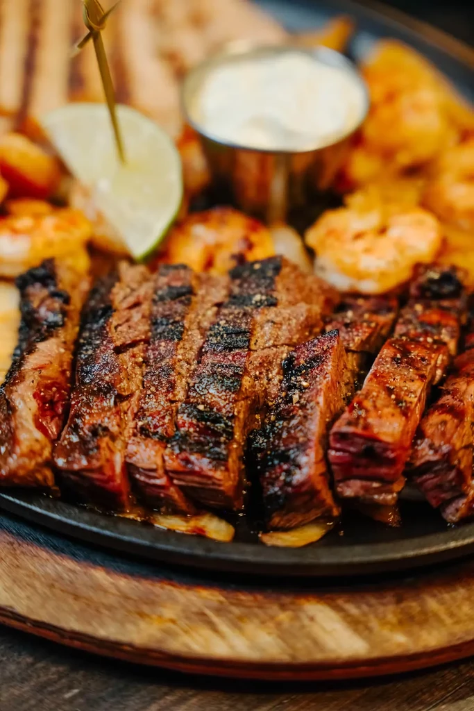 Sizzling platter with sliced steak, shrimp, lime, and white sauce.