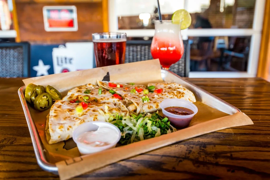 Quesadilla, lettuce, sauces, jalapeños, and drinks on a wooden table.