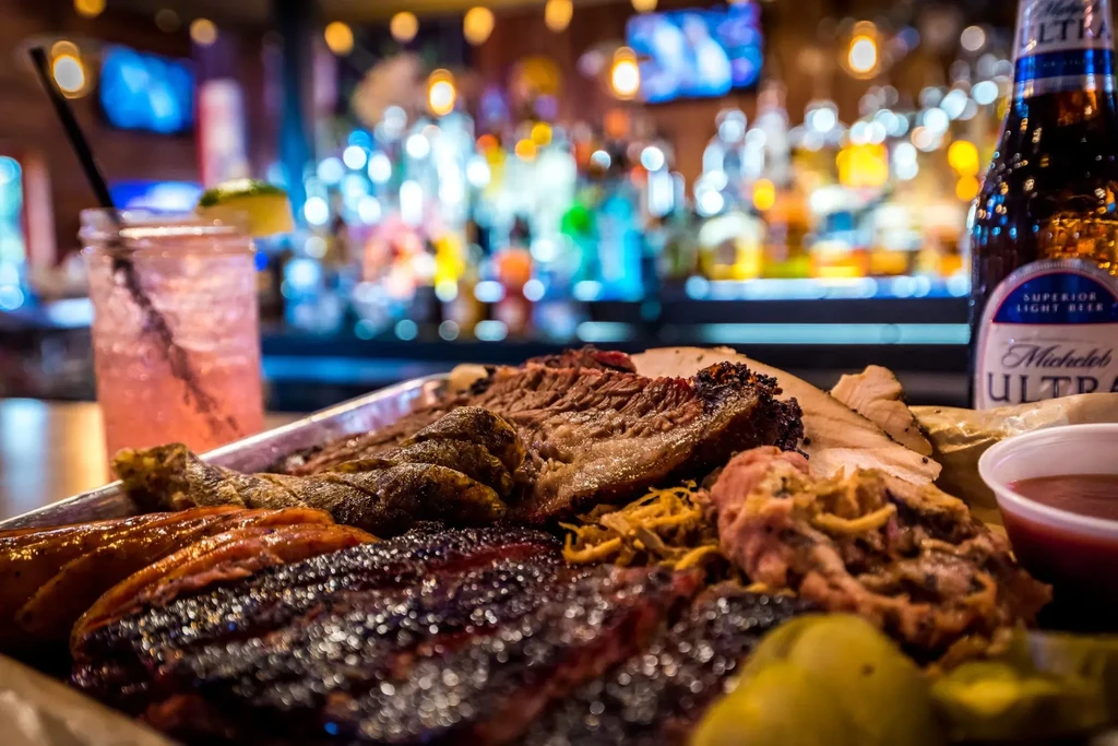 Close-up of a platter of barbecue meats with drinks and blurred background.