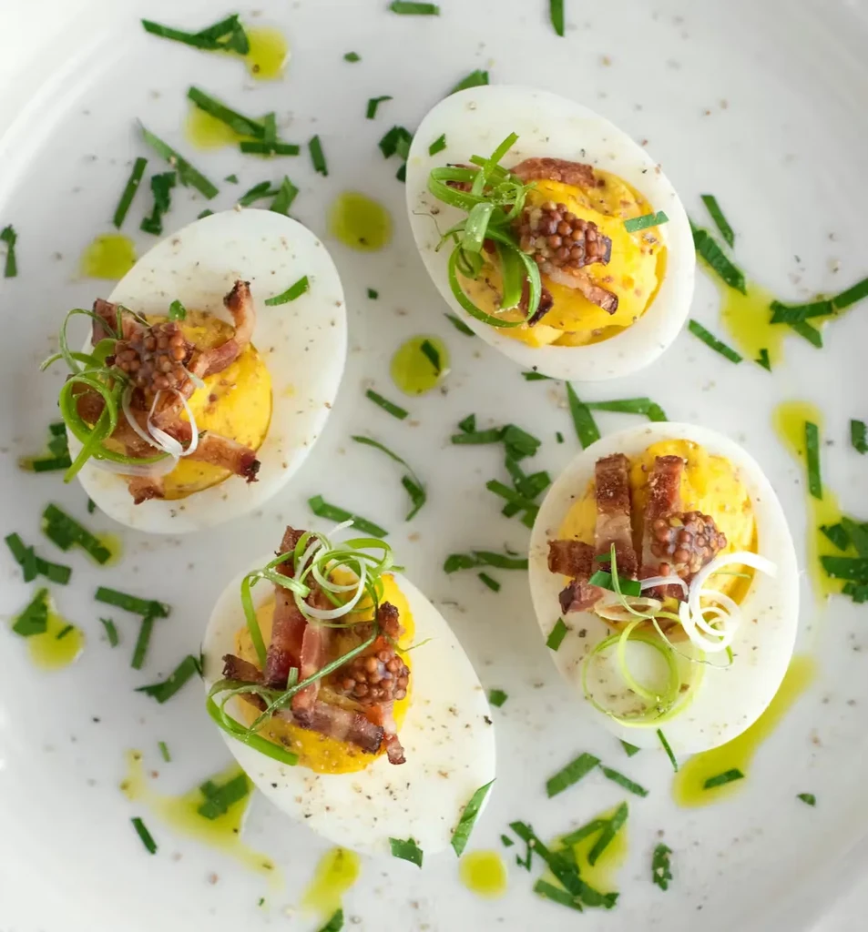 Four deviled eggs on a white plate, garnished with bacon and herbs.