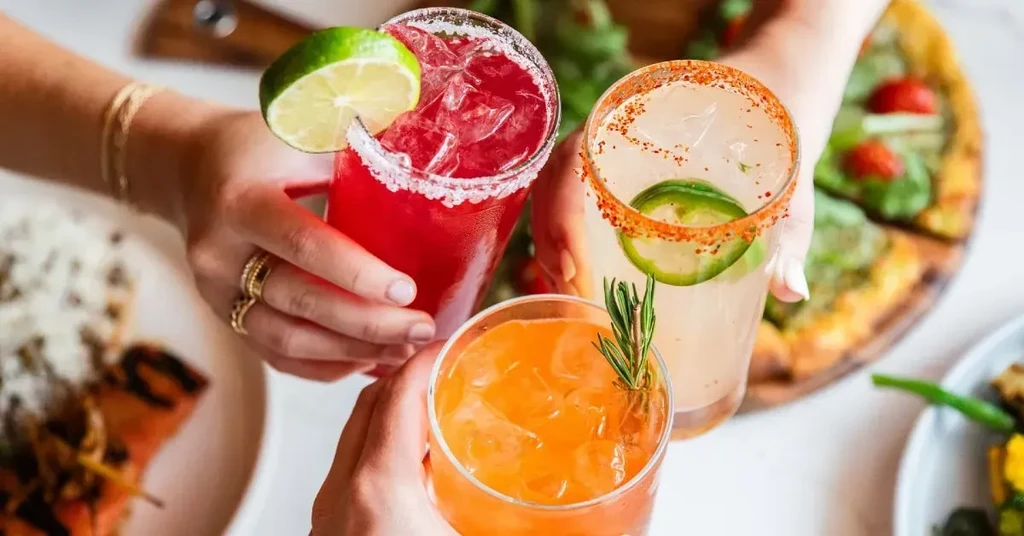 Three hands holding colorful cocktails in a toast over food.