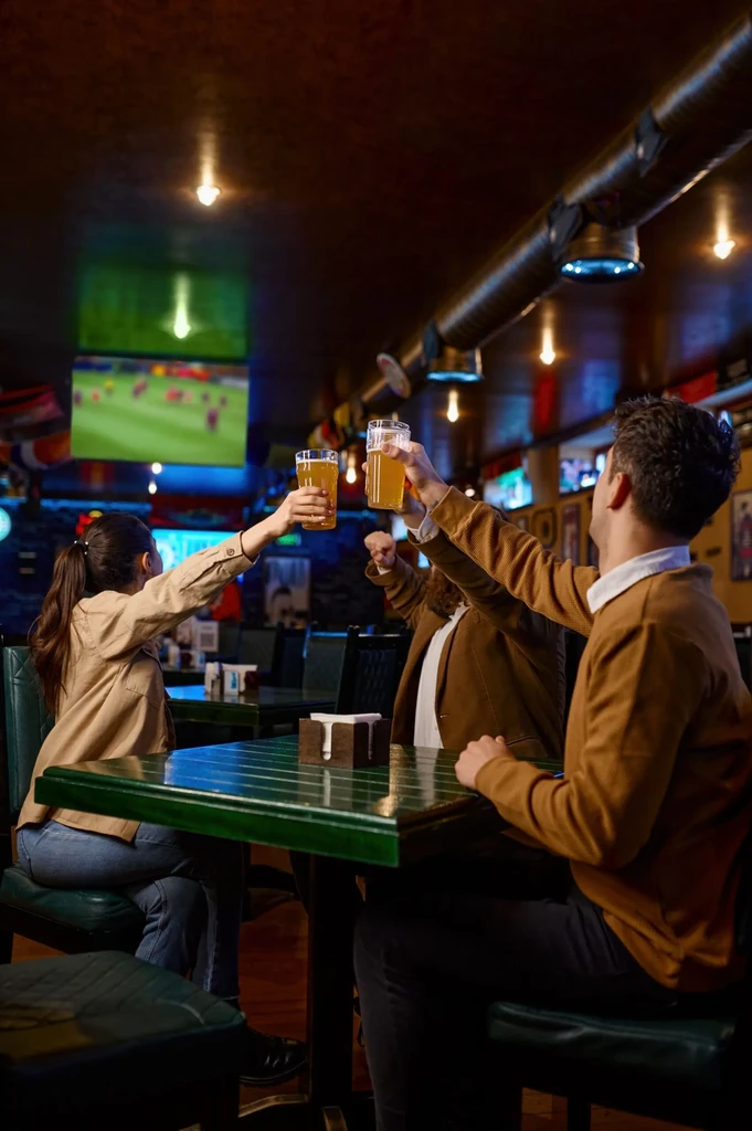 Three people toast with beer in a lively, warm-lit sports bar.