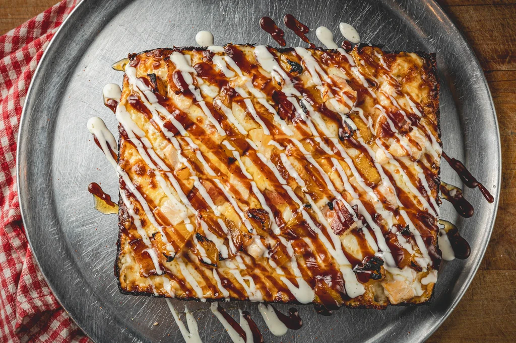 Rectangular pizza with BBQ and creamy sauce drizzles on a metal pan.