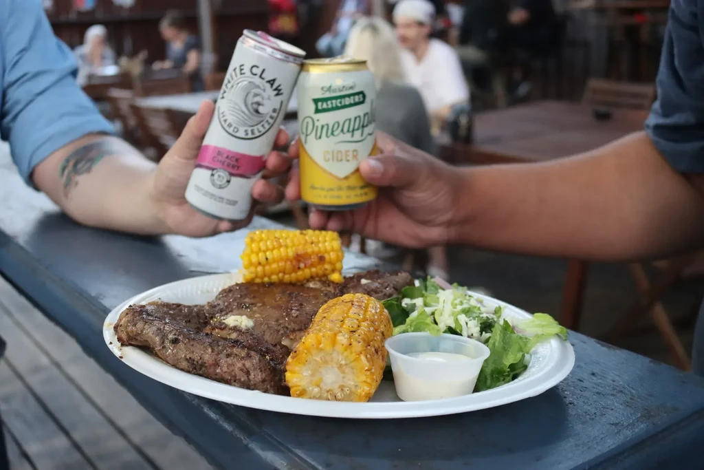Two hands hold a White Claw and cider above a meal.