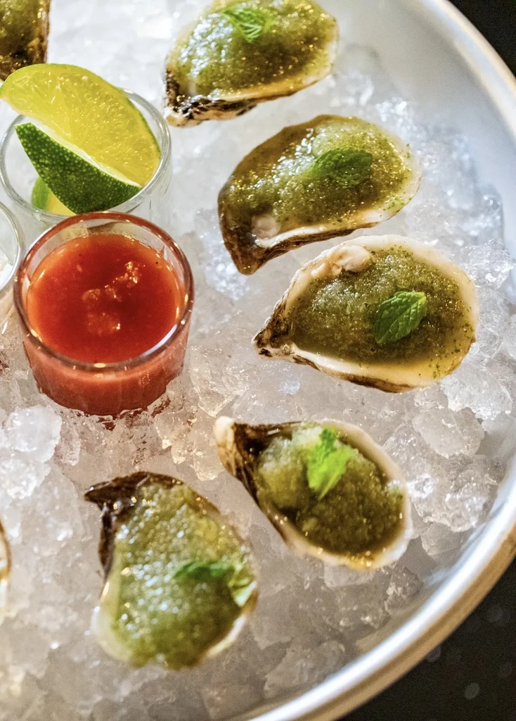 Platter of six oysters on ice with red sauce and lime wedges.