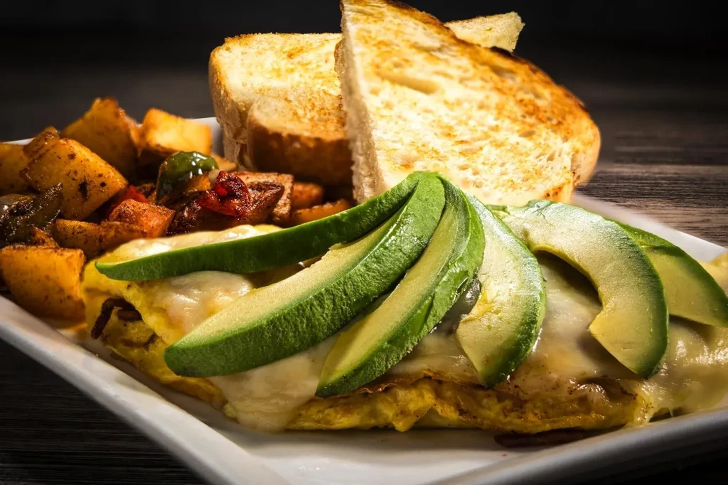 Breakfast plate with omelet, melted cheese, avocado, roasted potatoes, and toast.