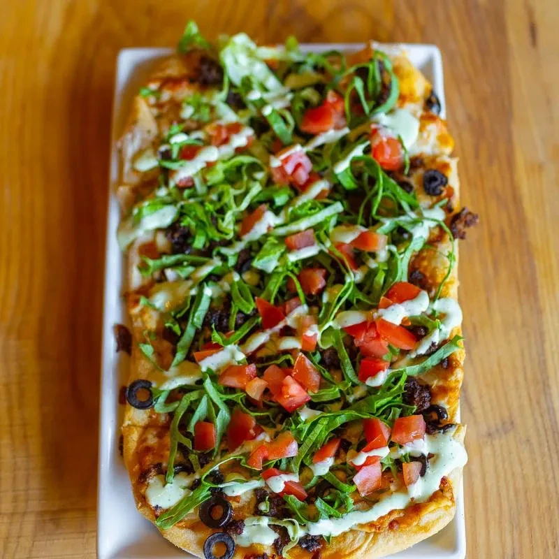 Rectangular flatbread with lettuce, tomatoes, olives, ground meat, and green sauce.