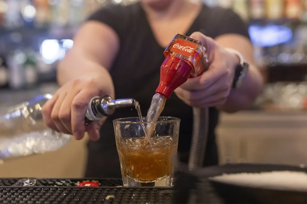 Bartender's hands pouring clear liquid and Coca-Cola into an iced glass.