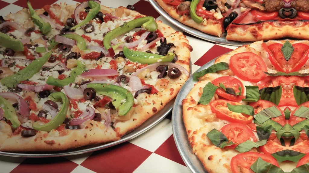 Overhead view of three pizzas with diverse vegetable toppings on checkered tablecloth.