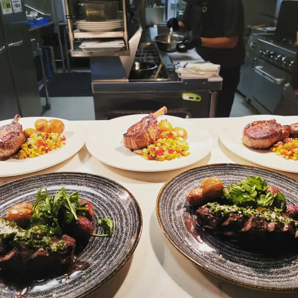 Professionally plated steak and pork chops on a counter; chef cooks in background.