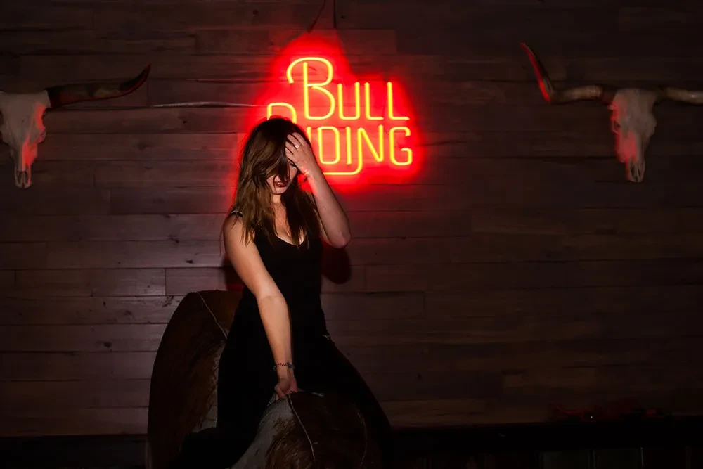 Woman on mechanical bull, red neon "BULL RIDING" sign behind her.