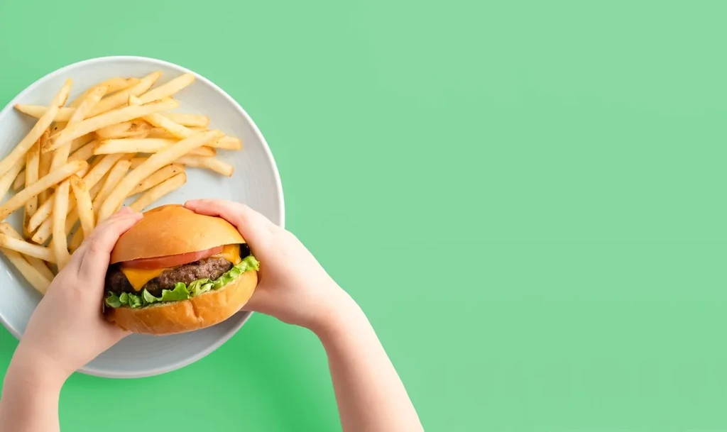 Top-down, child's hands hold cheeseburger and fries on a plate.
