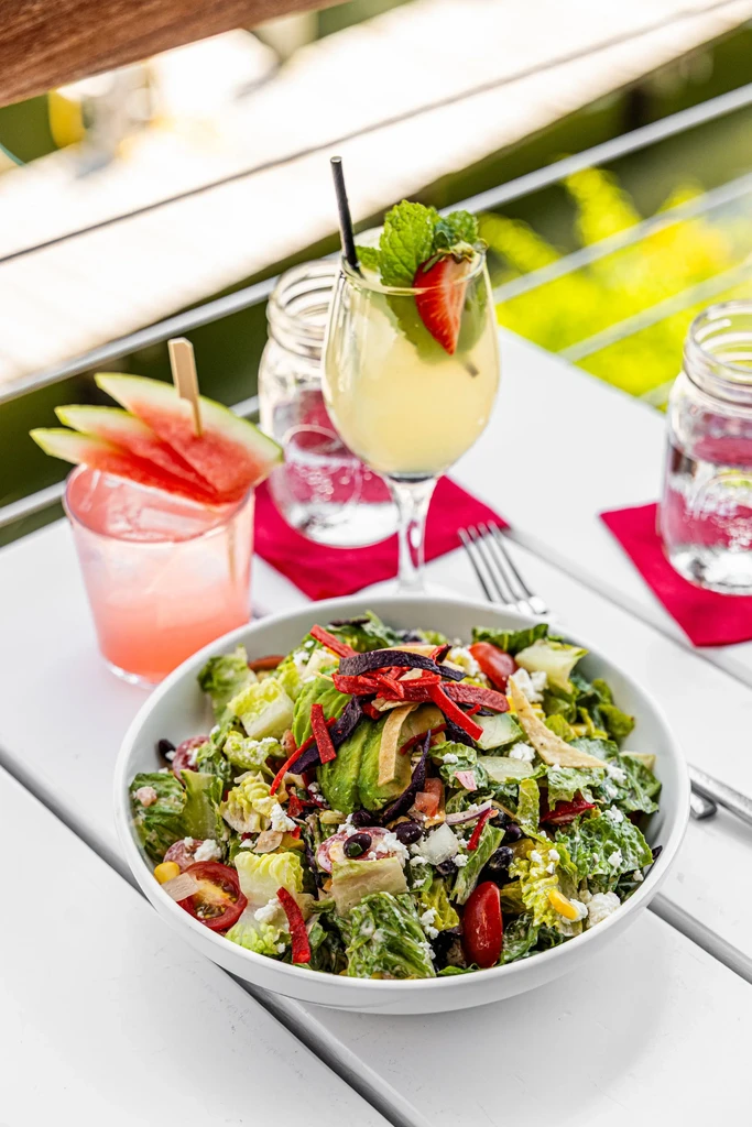 Colorful salad, two cocktails, and water on a slatted outdoor table.
