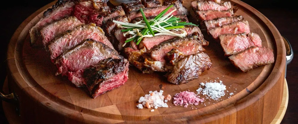 Sliced medium-rare steak, garnished, with three gourmet salts on a board.