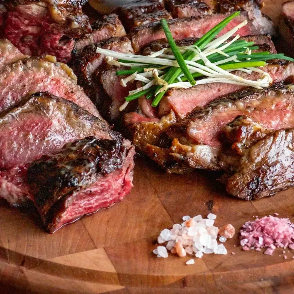 Sliced medium-rare steak on wood board with chives, mushrooms, and two salts.