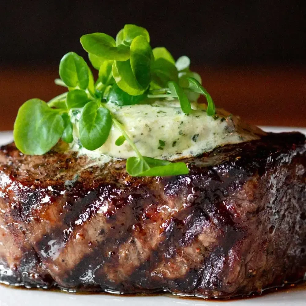 Thick seared steak with grill marks, melting herb butter, and green garnish.