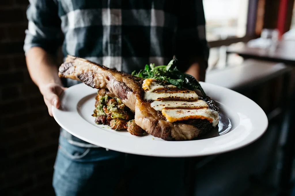 Artfully plated bone-in meat topped with charred cheese and roasted vegetables.