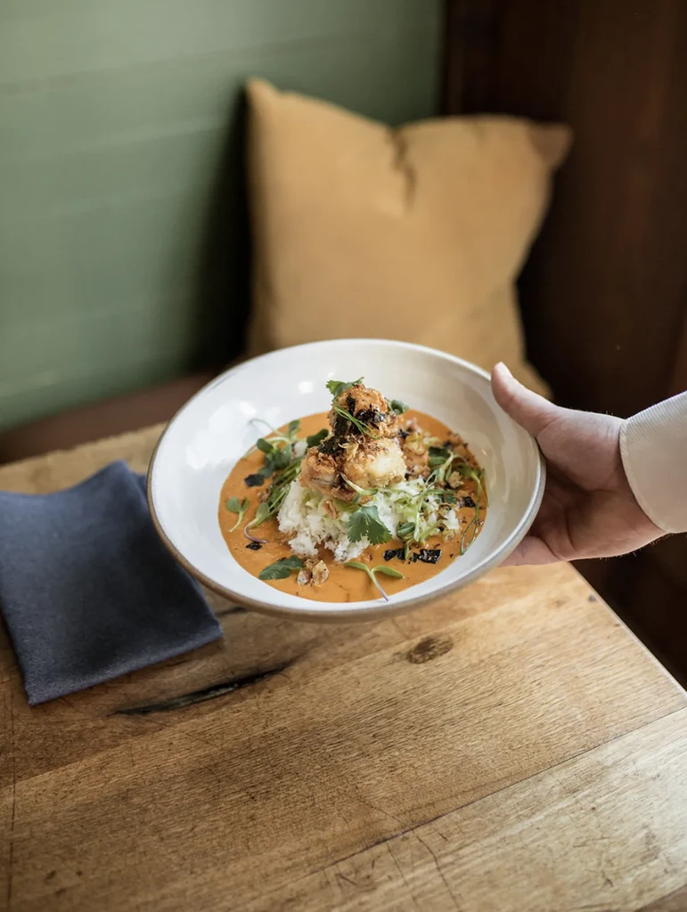 Hand holds plated curry with rice, crispy protein, fresh herbs.