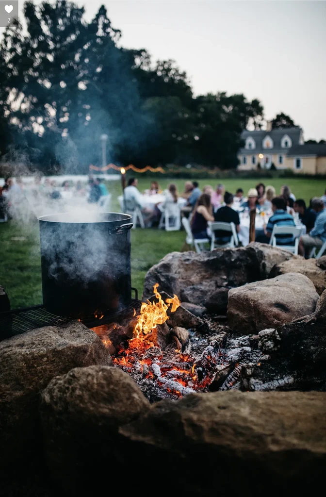 Large pot steaming over fire pit, blurred people at evening event.