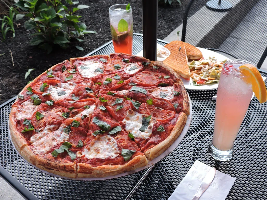 Outdoor dining scene with sliced pizza, grilled sandwich, pasta salad, and two drinks.