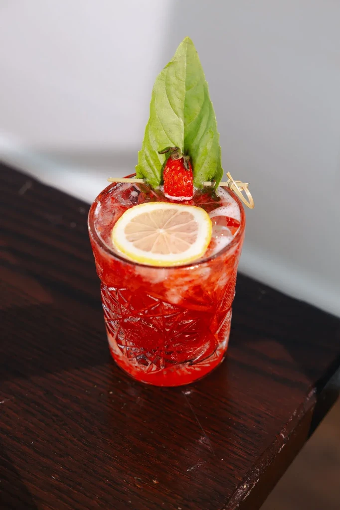 Red cocktail in textured glass, garnished with strawberry, lemon, leaf.