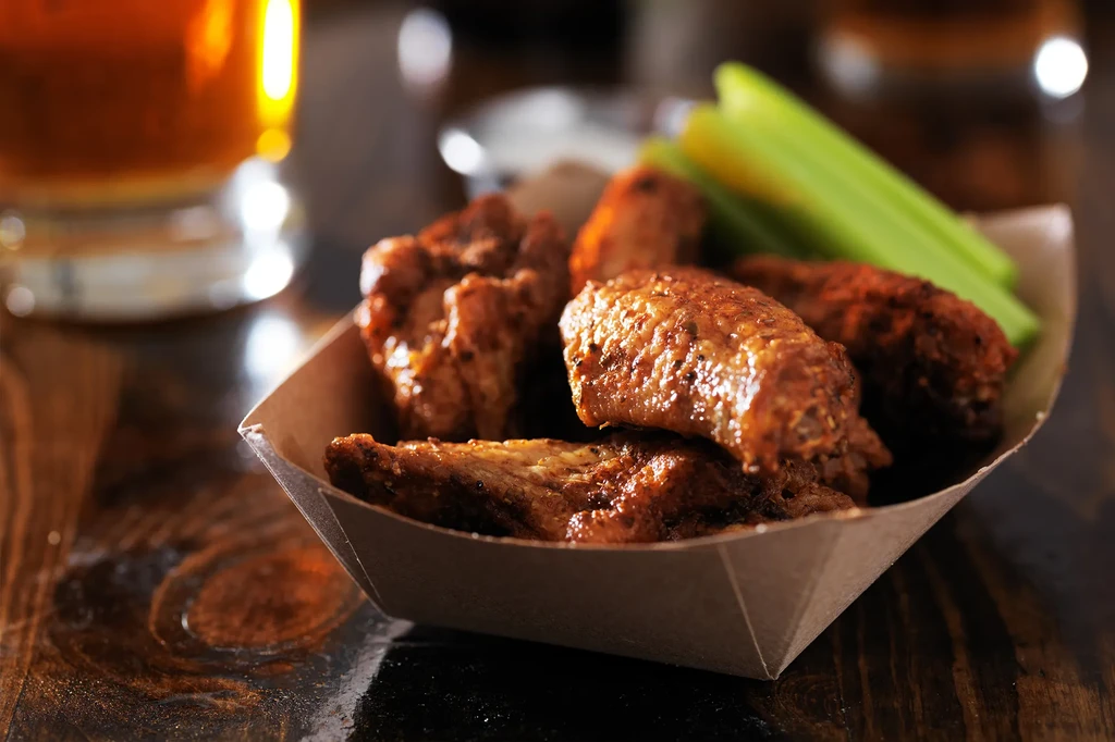 Crispy chicken wings with celery sticks in a paper boat on wood.