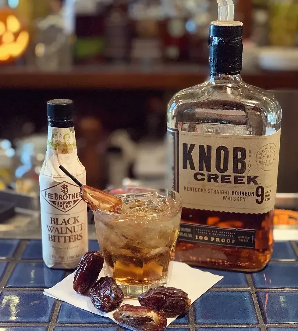 Bourbon cocktail, dried dates, bitters, and whiskey bottles on a bar.