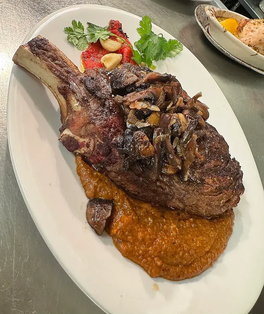 Large Tomahawk steak with mushrooms, sauce, roasted peppers, and herbs.