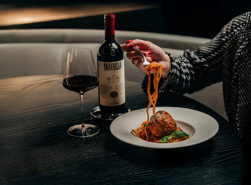 Spaghetti with meatball, red wine, and a person's hand with fork.