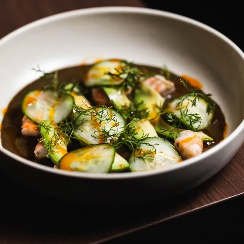 A white bowl with sliced cucumbers, avocado, and shrimp in sauce.
