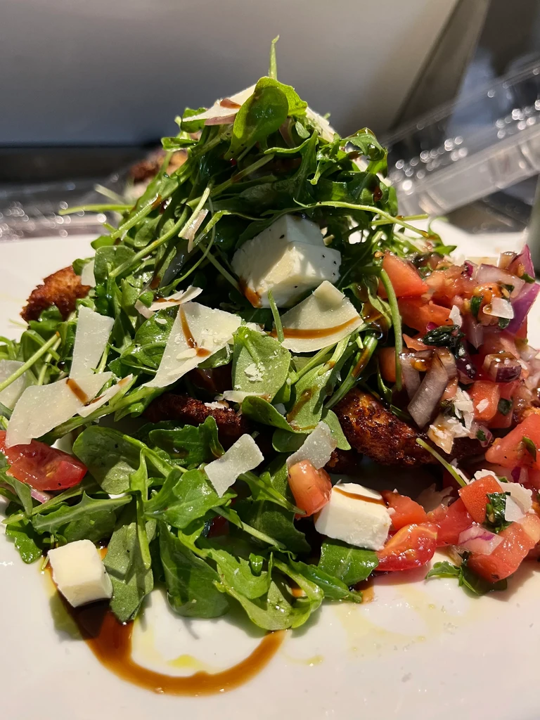 Arugula salad with shaved cheese, tomatoes, and balsamic glaze on a white plate.
