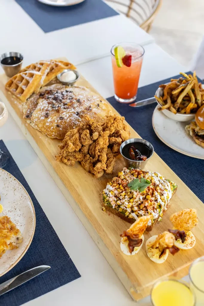 Wooden board with waffles, pancakes, fried chicken, avocado toast, deviled eggs.