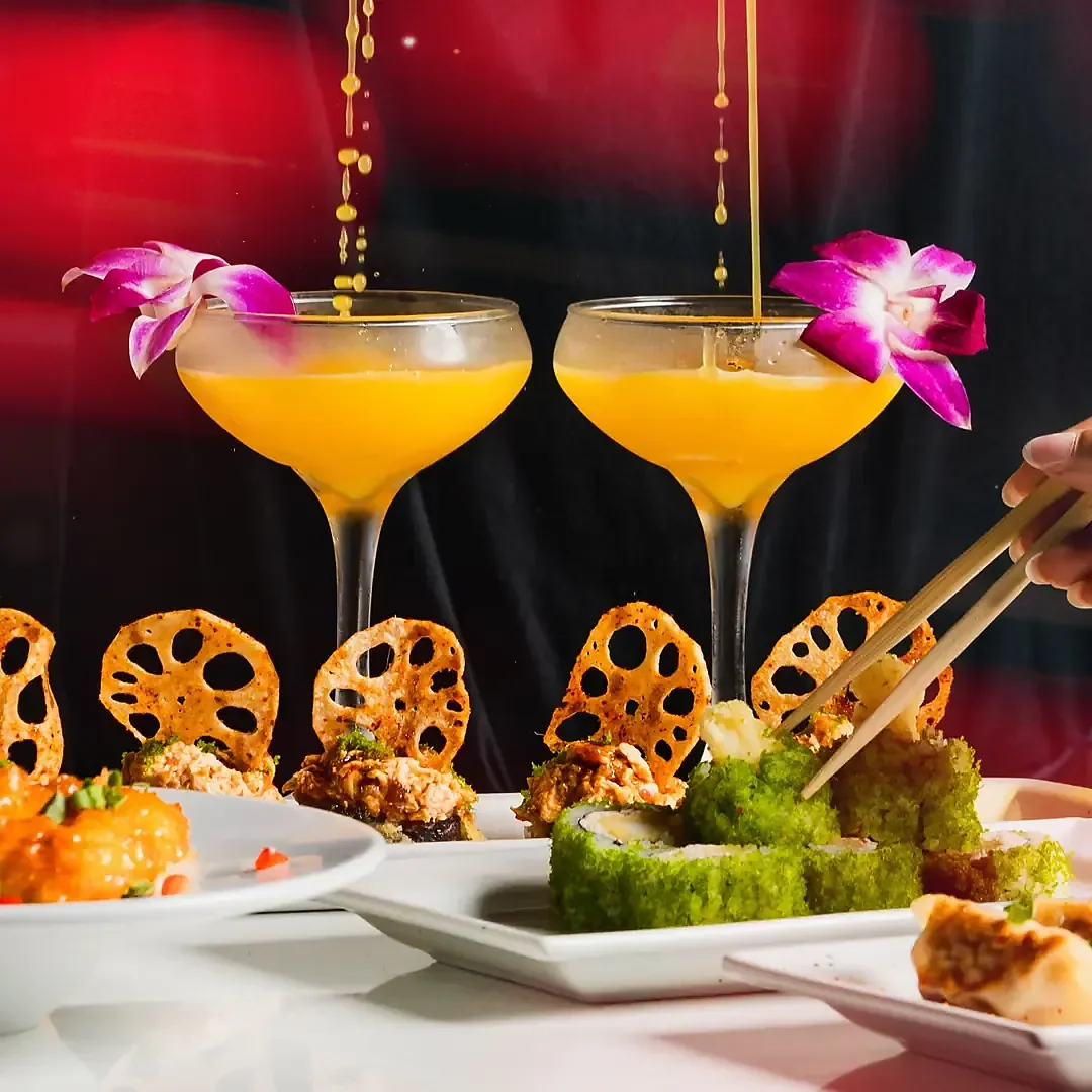 Two orange cocktails with orchid garnish beside sushi rolls and lotus chips