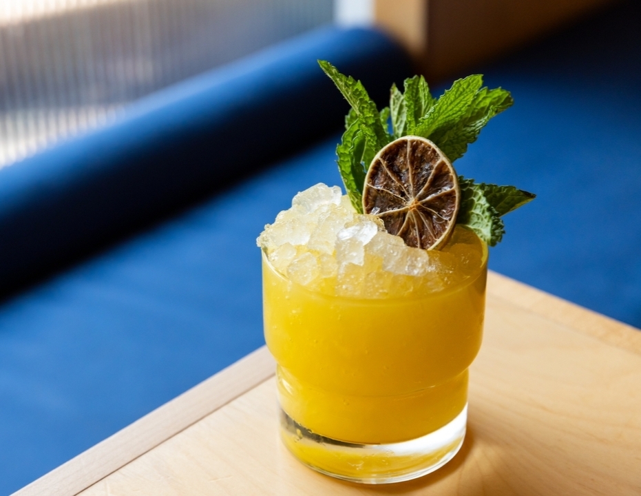 Yellow drink in glass, topped with crushed ice, mint, dried lime.