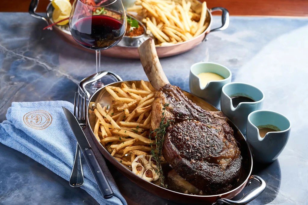 Tomahawk steak with fries and sauces on a marble tabletop.