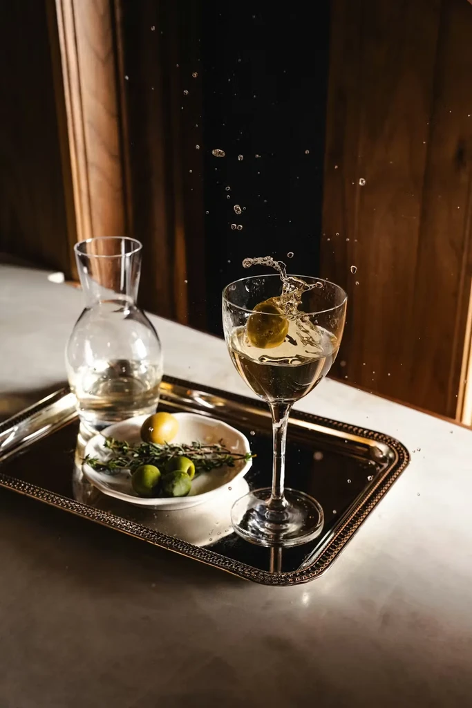 Martini glass with splashing olive, decanter, and olives on a silver tray.