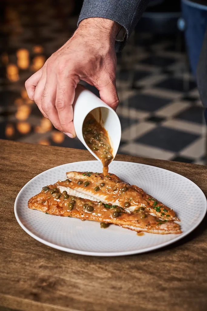 Pan-fried fish fillets with caper sauce are drizzled on a white plate.