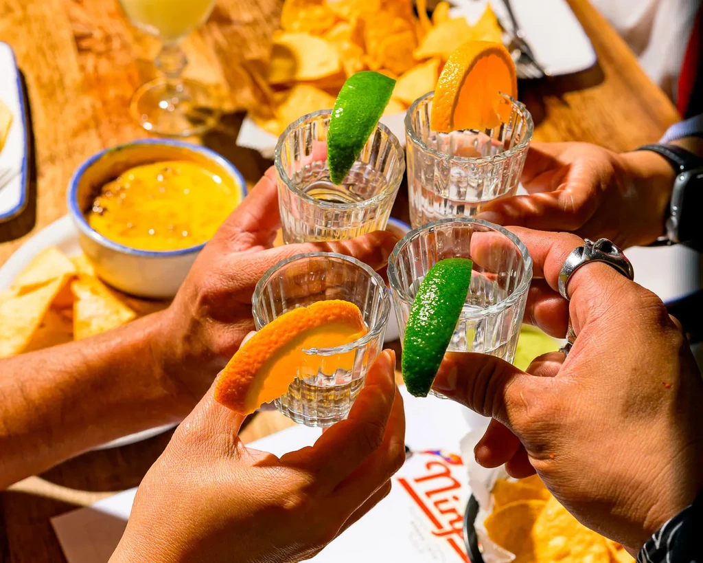Four hands toast shot glasses garnished with lime and orange.