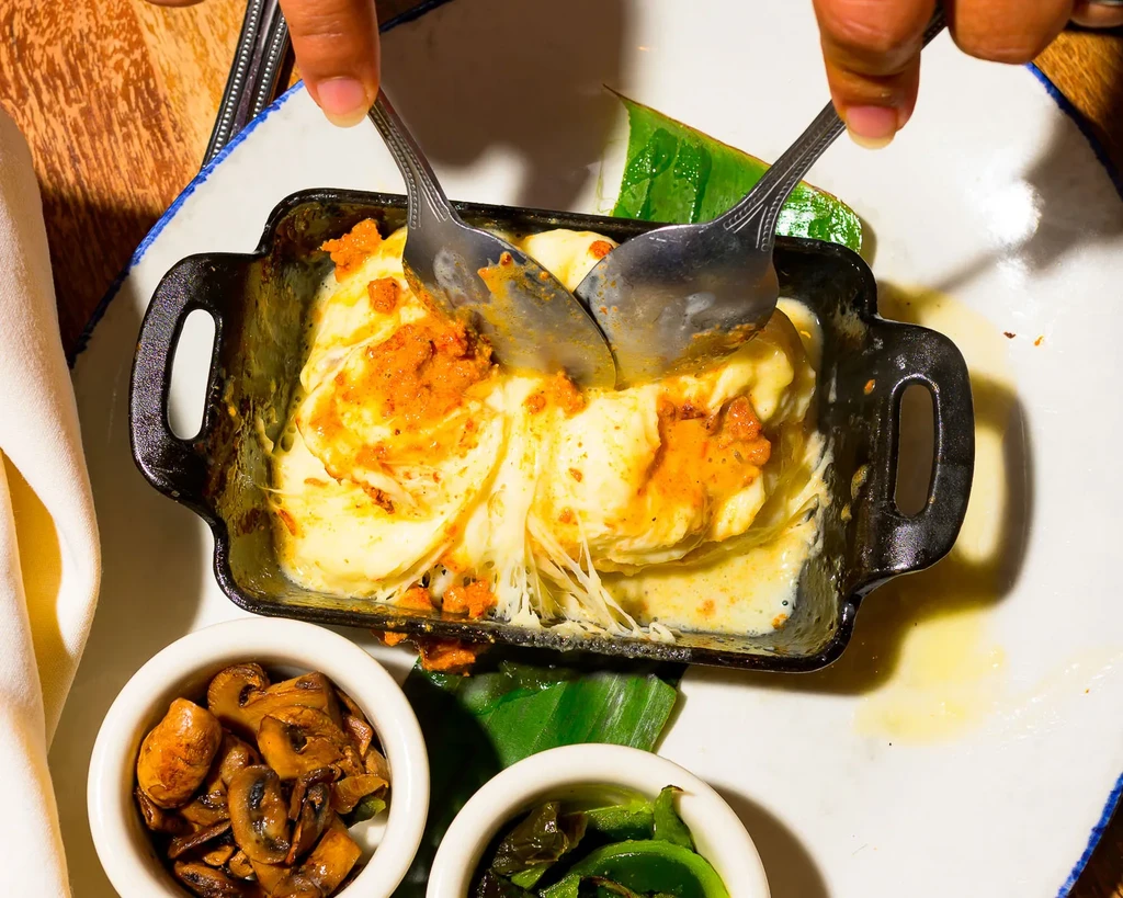 Hands scooping melted cheesy dish from cast iron skillet with sides.