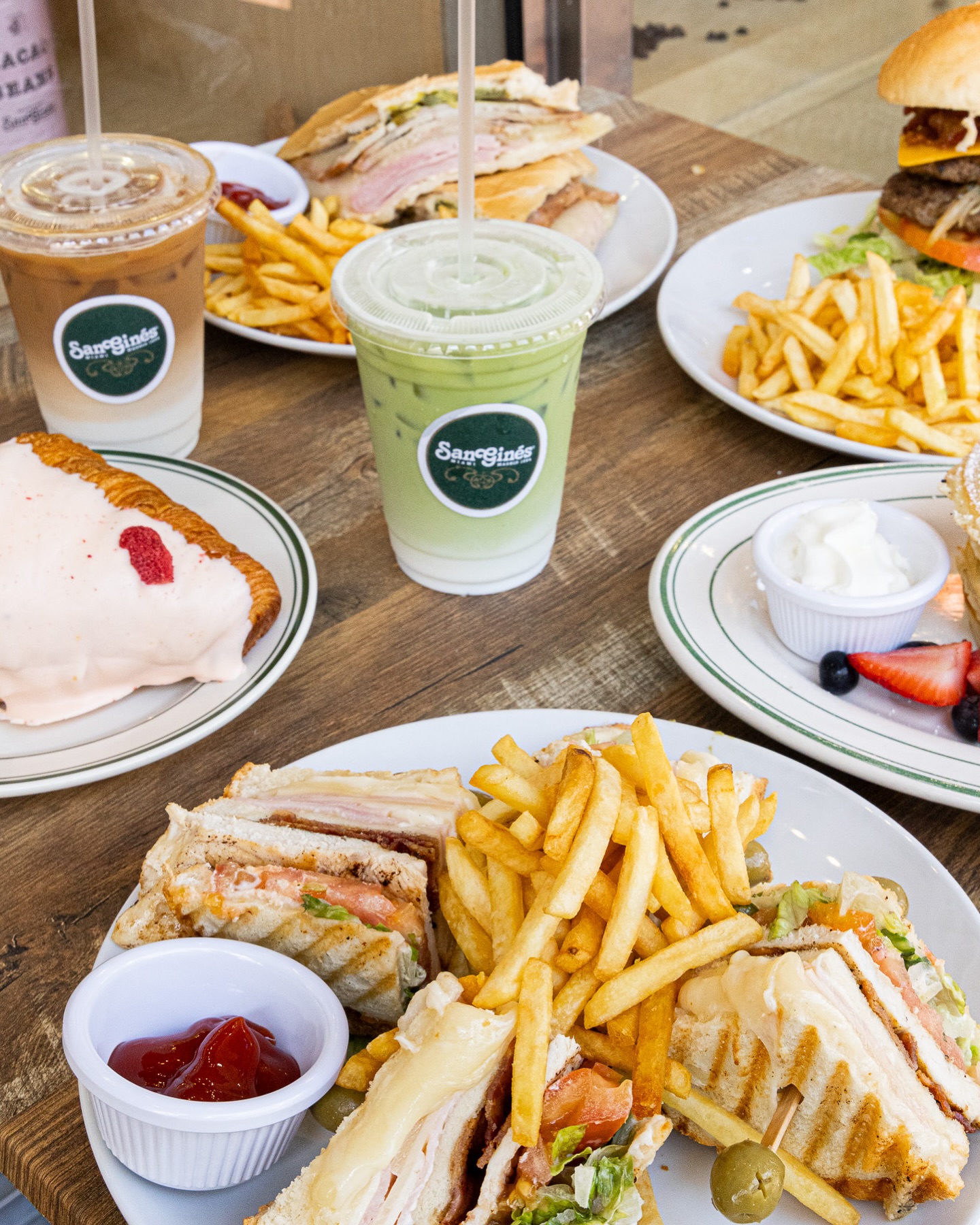 Various sandwiches, burgers, fries, drinks, and a frosted croissant on a table.