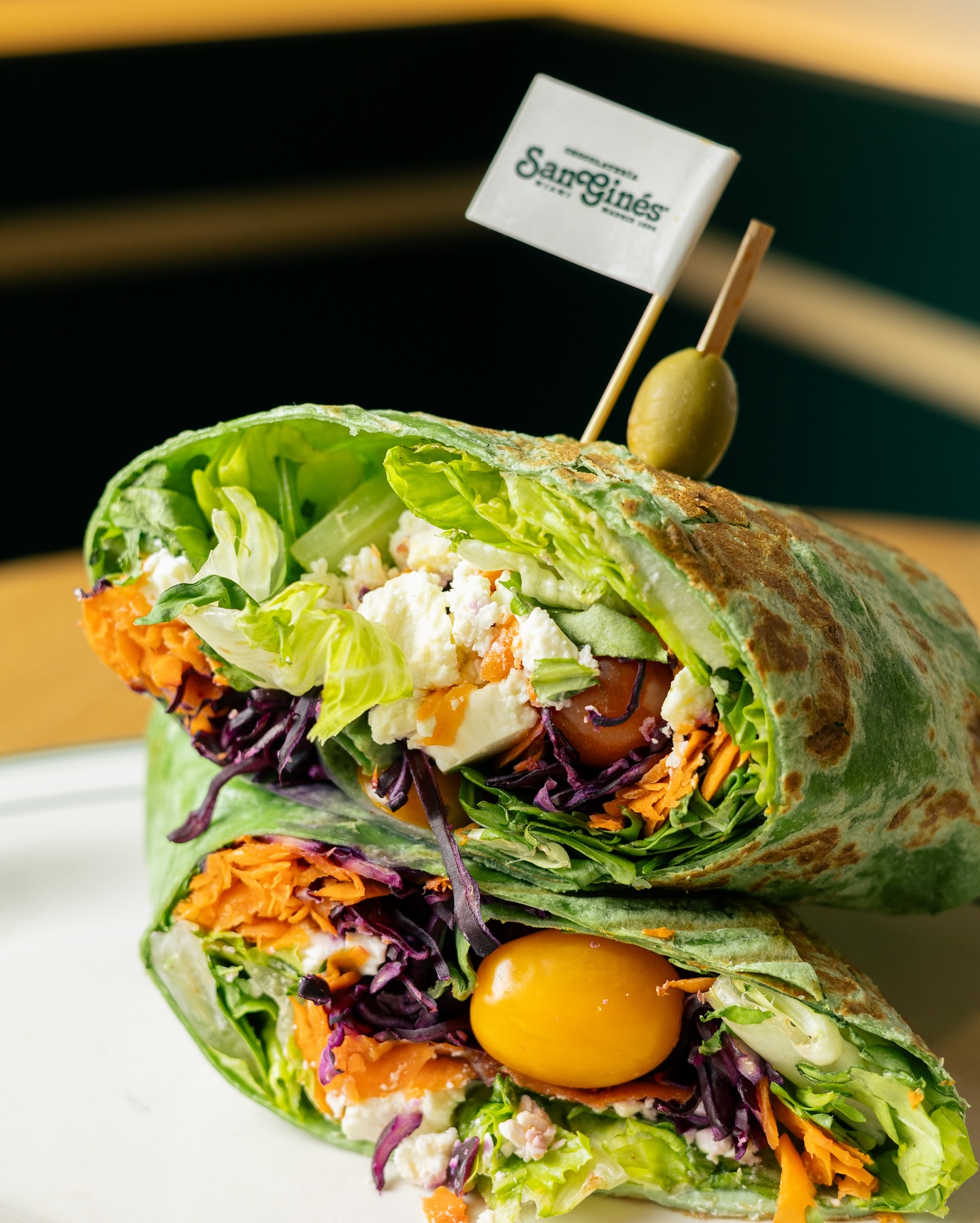 Two halves of a green wrap with vegetables, cheese, and San Ginés flag.