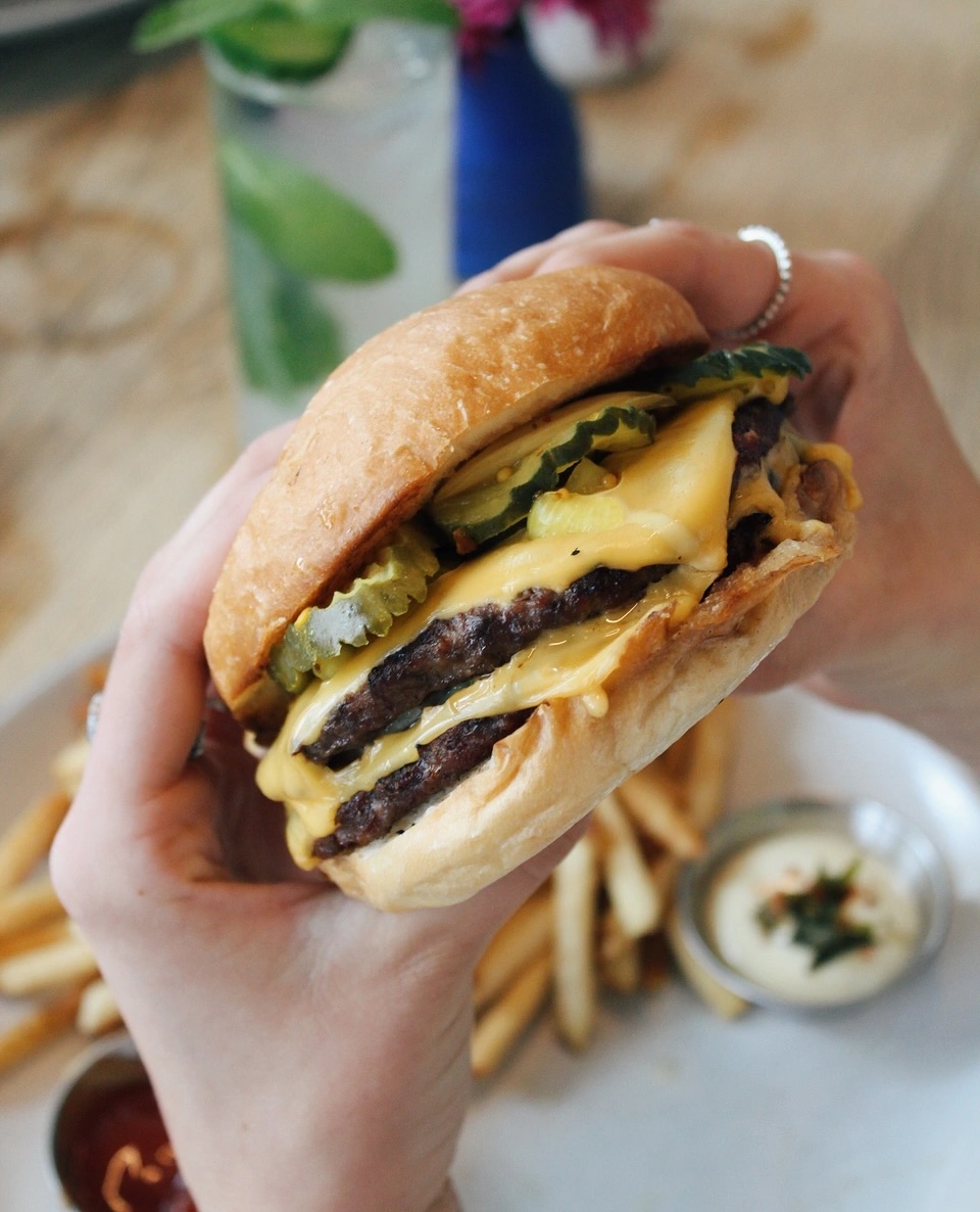 A person holds a double cheeseburger with pickles and melted cheese.