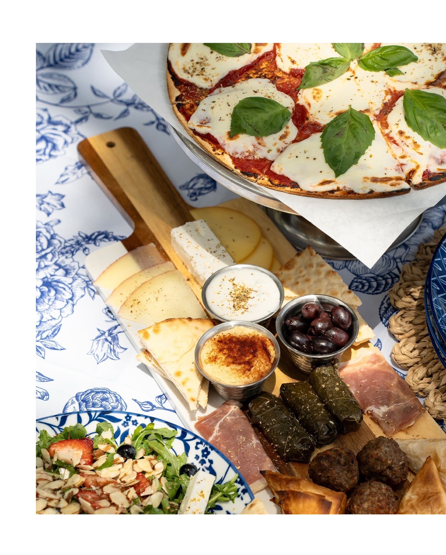 Margherita pizza, charcuterie board, and salad on a blue floral tablecloth.