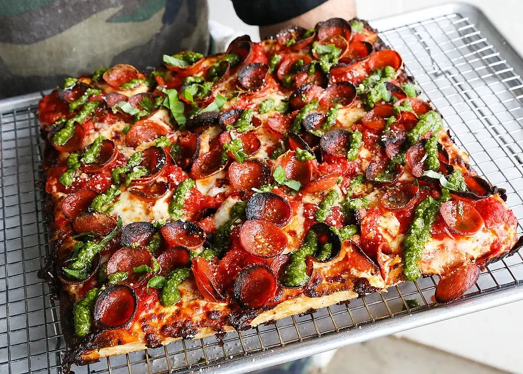 Square pan pizza topped with curled pepperoni and green pesto drizzle