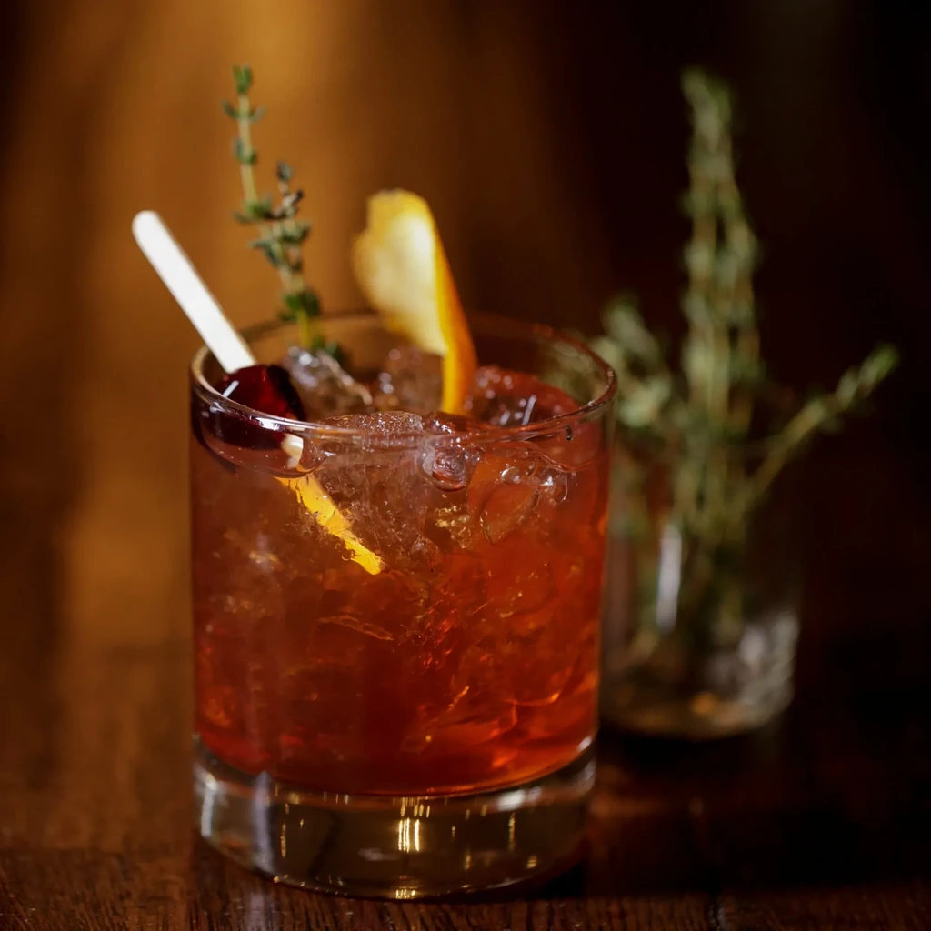 Amber cocktail in a rocks glass, garnished with herbs and orange.