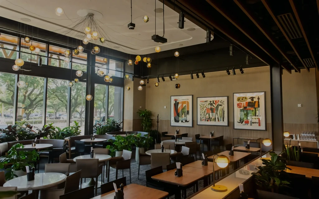 Modern restaurant interior with tables, chairs, plants, and large windows.
