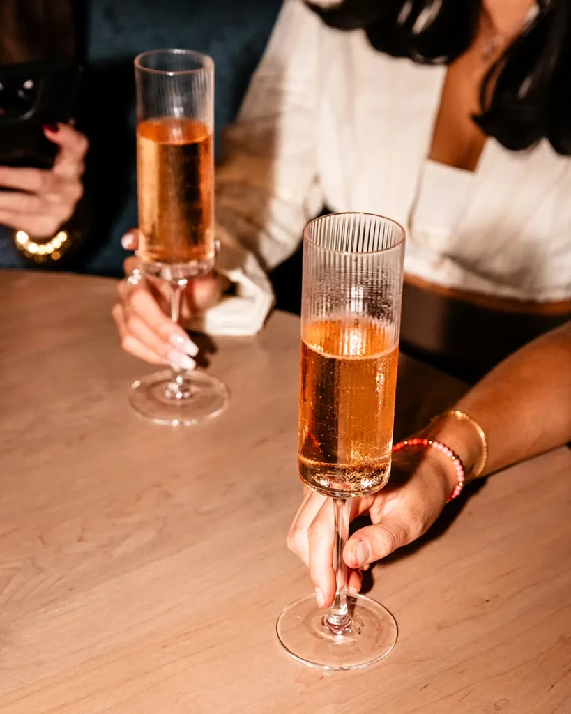 Two hands hold fluted glasses filled with a sparkling amber beverage.